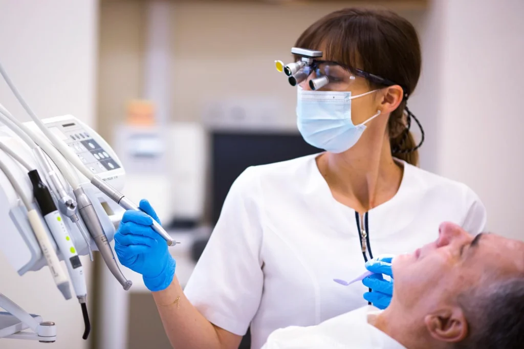 Dentystka ubrana w biały fartuch, maseczkę ochronną i specjalne okulary z lupą, trzyma w ręku narzędzie dentystyczne, przygotowując się do zabiegu. Pacjent leży na fotelu dentystycznym w nowoczesnym gabinecie, na pierwszym planie widoczne są urządzenia stomatologiczne.